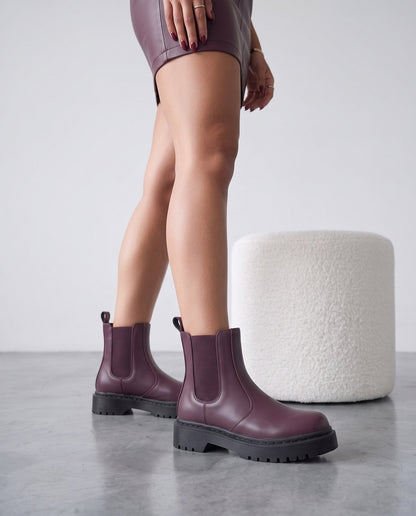 Dettaglio ravvicinato di stivaletti Chelsea bordeaux con suola nera, gambe della modella in abito di pelle bordeaux, pouf bianco sullo sfondo.
