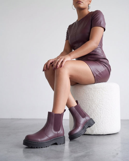 Modella seduta su pouf bianco, stivaletti chelsea bordeaux in primo piano, gambe incrociate alle caviglie