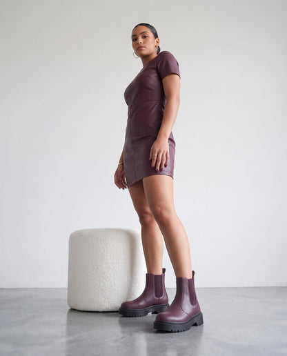 Modella in abito di pelle bordeaux e stivaletti chelsea bordeaux con suola nera, in piedi accanto a pouf bianco