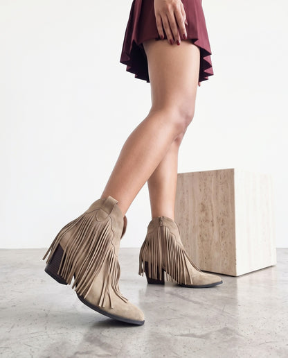 Gambe di modella con stivaletti khaki con frange, scatto ravvicinato dal basso accanto a cubo in travertino.