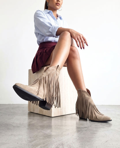 Modella seduta su cubo in travertino, gambe accavallate, stivaletti khaki con frange in camoscio