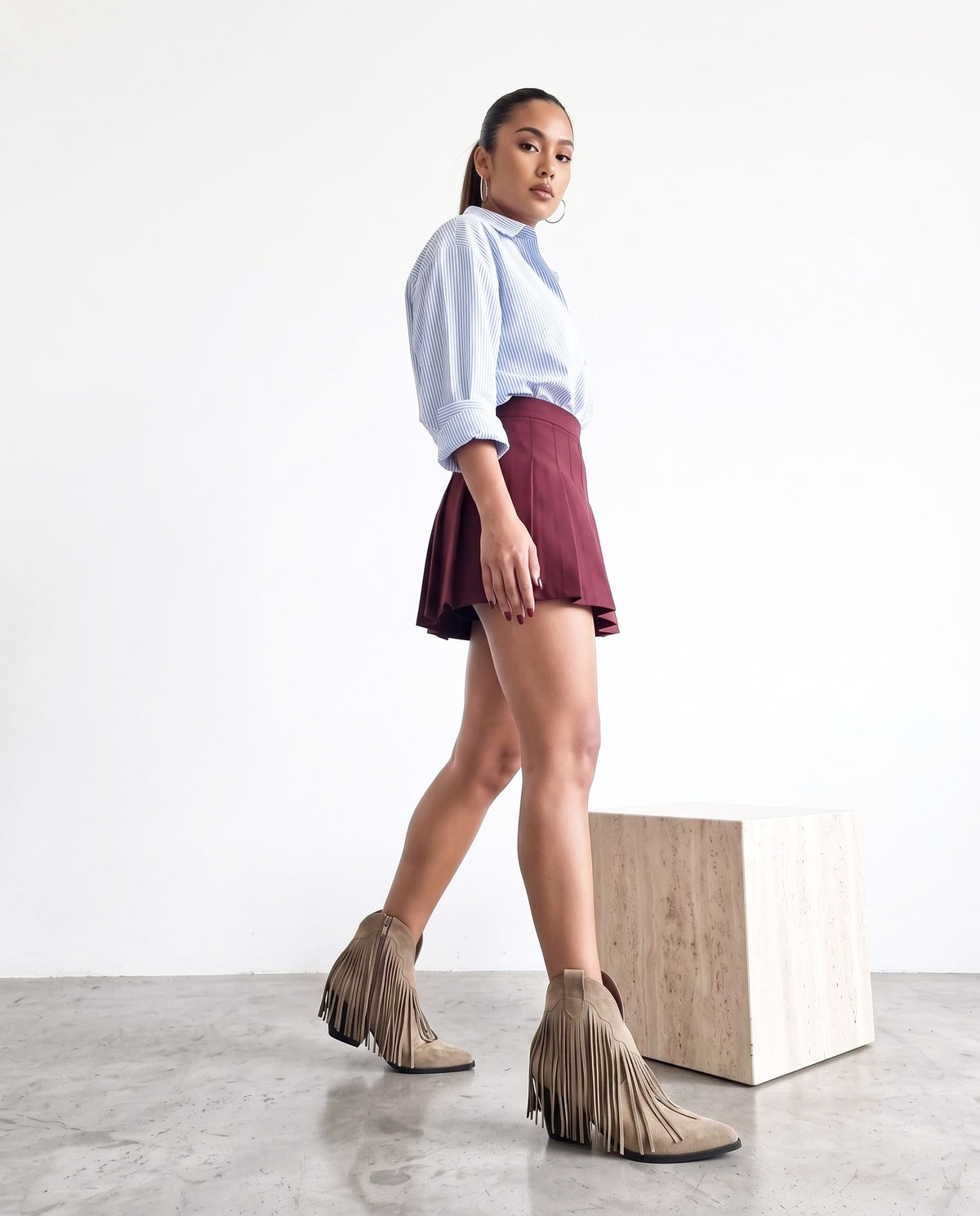 Modella di profilo con stivaletti khaki a frange, camicia a righe e minigonna bordeaux, in cammino