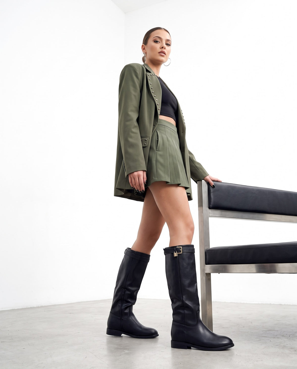 Modella con stivali alti neri, blazer e minigonna verde militare, accanto a una panca con seduta in pelle nera