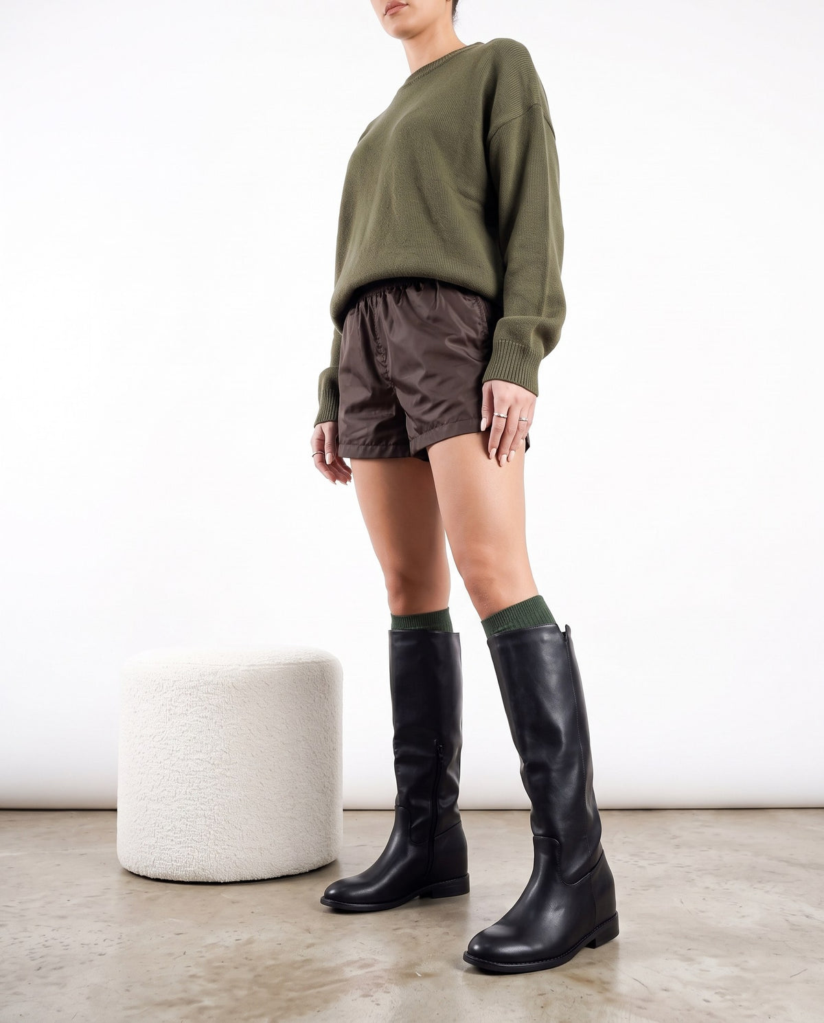 Modella in piedi con stivali neri al ginocchio, maglia verde oliva e shorts marroni, accanto a pouf bianco