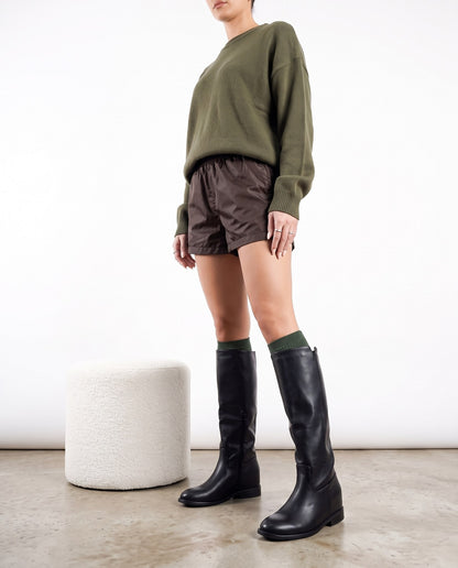 Modella in piedi con stivali neri al ginocchio, maglia verde oliva e shorts marroni, accanto a pouf bianco