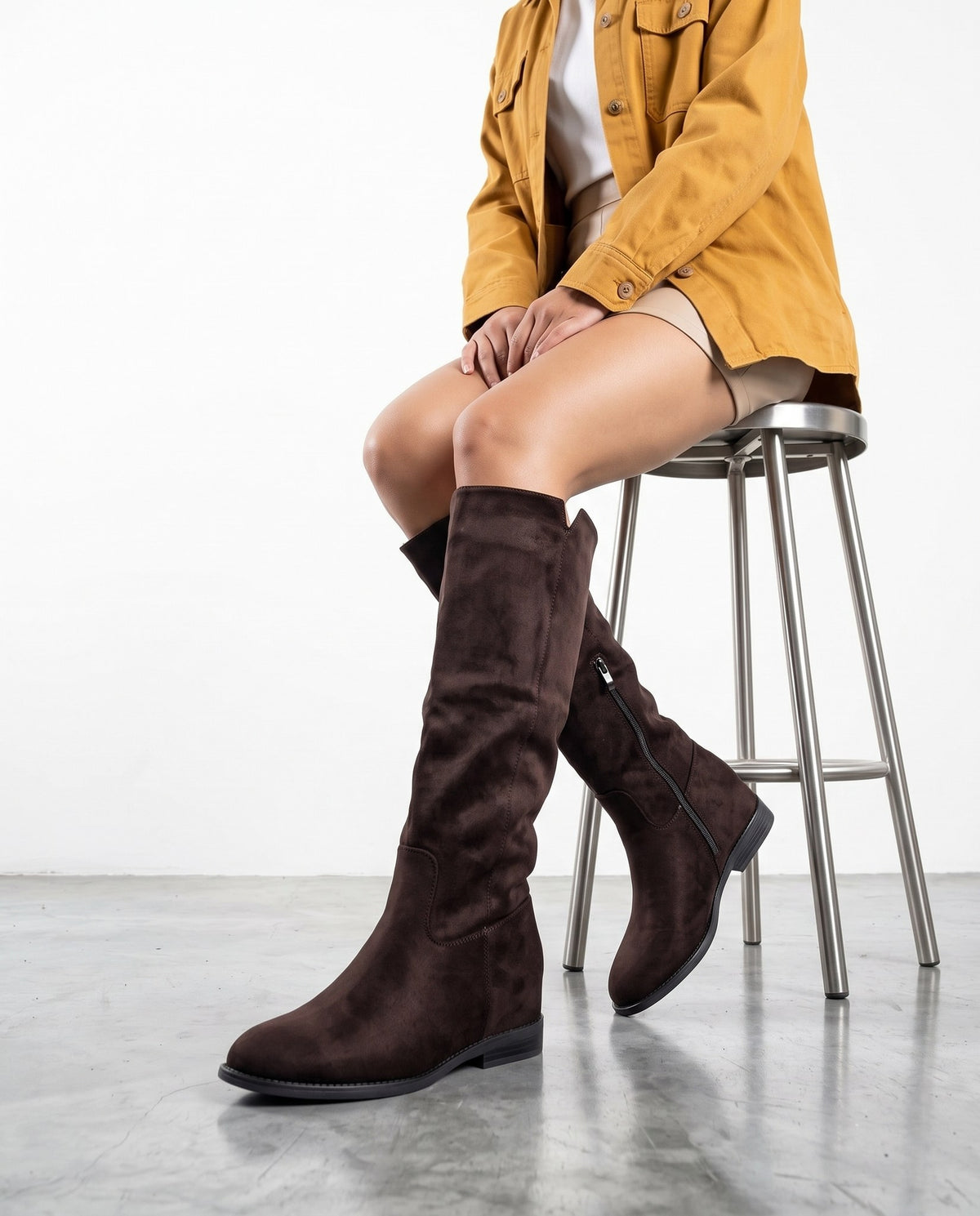 Modella seduta su sgabello cromato, stivali alti marrone scuro in camoscio con cerniera laterale, gambe accavallate