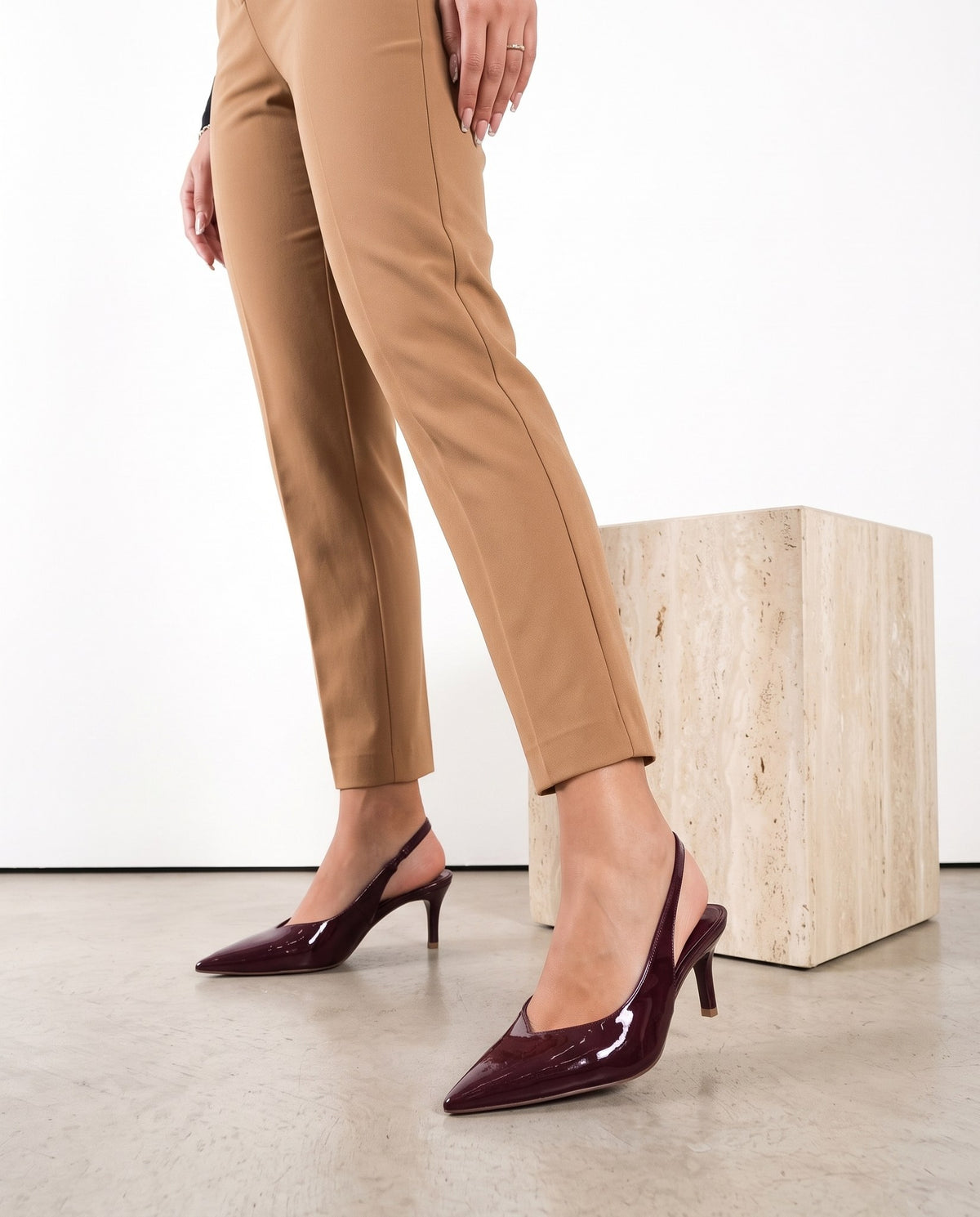 Scarpe décolleté slingback bordeaux lucide su gambe in pantaloni cammello, cubo in travertino sullo sfondo
