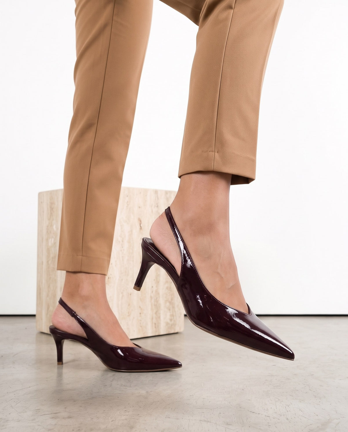 Dettaglio ravvicinato di scarpe décolleté slingback color vino lucido, gambe con pantaloni cammello, cubo in travertino sullo sfondo.