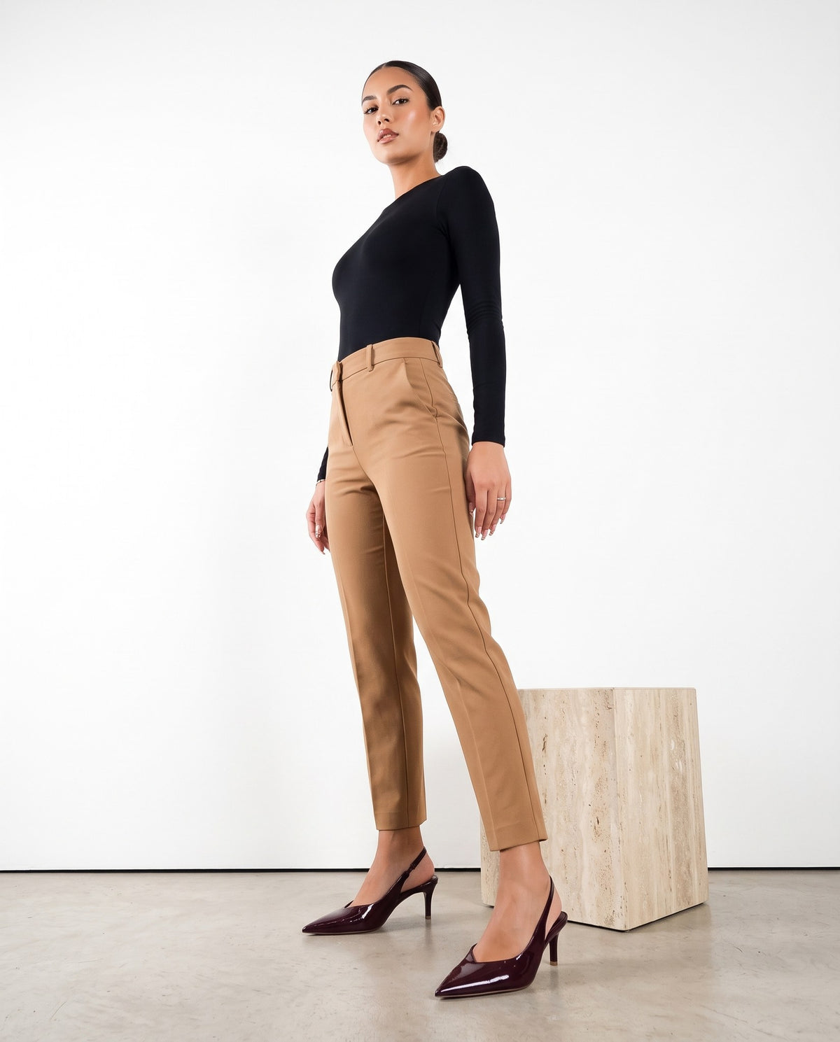 Modella in piedi con pantaloni cammello e décolleté slingback bordeaux lucide, accanto a cubo in travertino.
