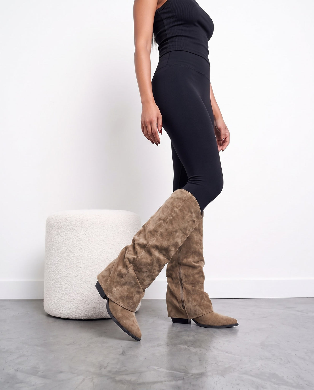 Gambe di modella di profilo con stivali camoscio khaki a gambale ripiegato, pouf bianco a sinistra