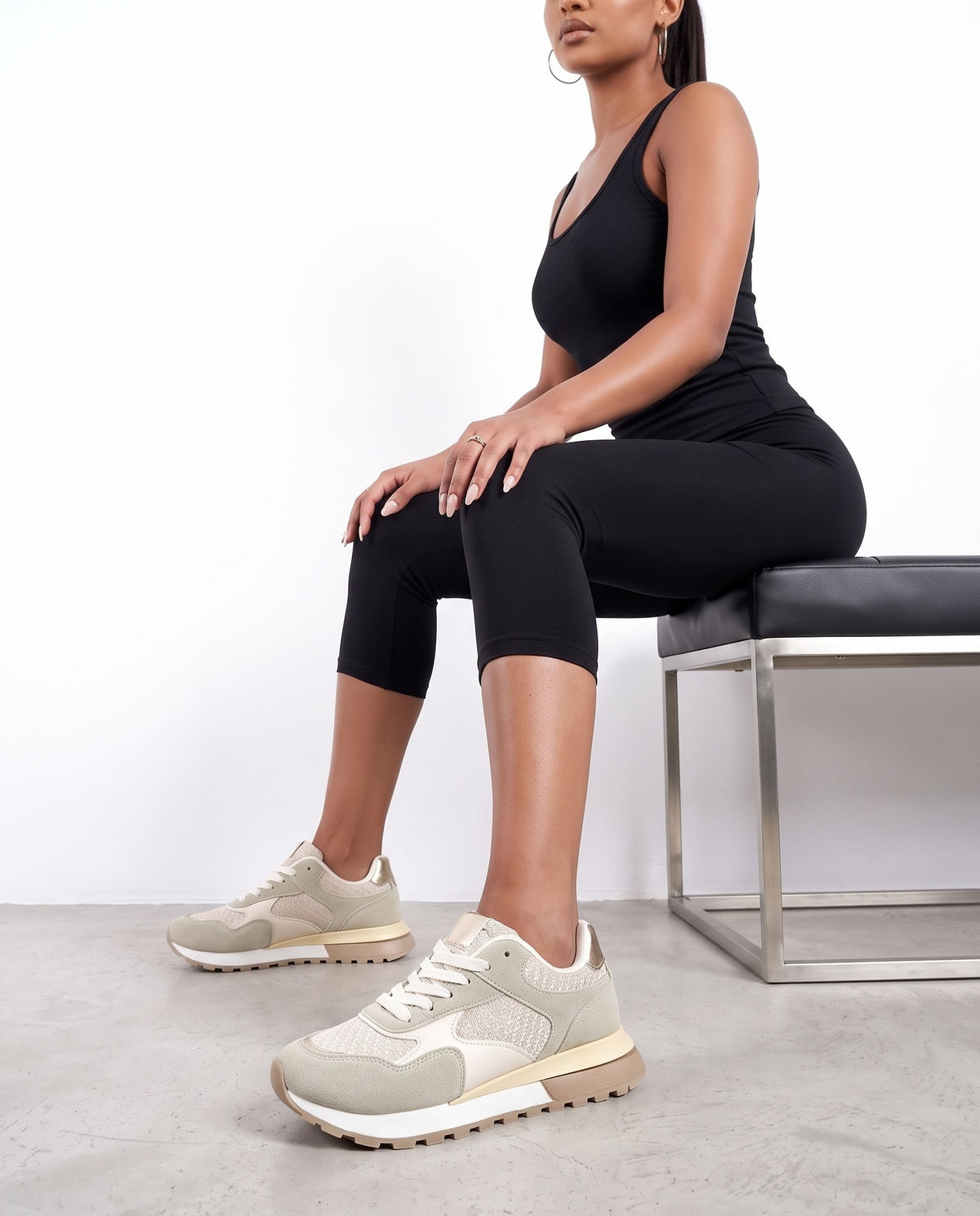 Modella seduta su panca, mostra sneakers beige da vicino, inquadratura dal busto in giù.