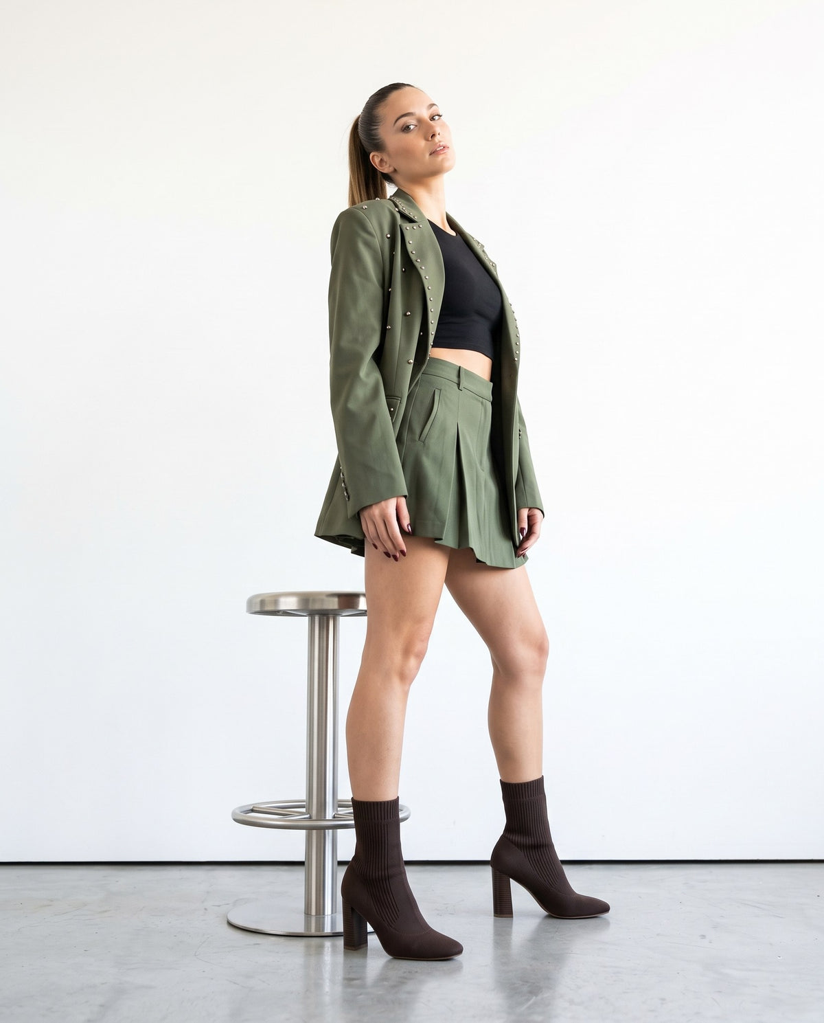 Modella in blazer e minigonna verde militare con stivaletti sock marroni, in piedi accanto a sgabello in metallo.