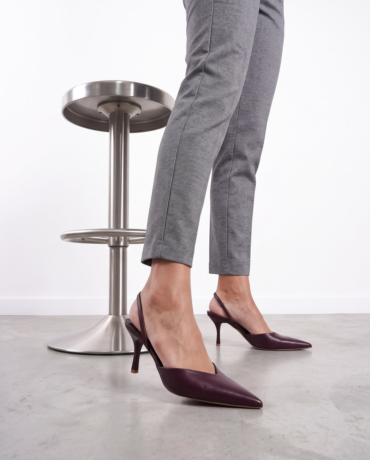 Gambe con pantaloni grigi e décolleté slingback color vino accanto a uno sgabello cromato.