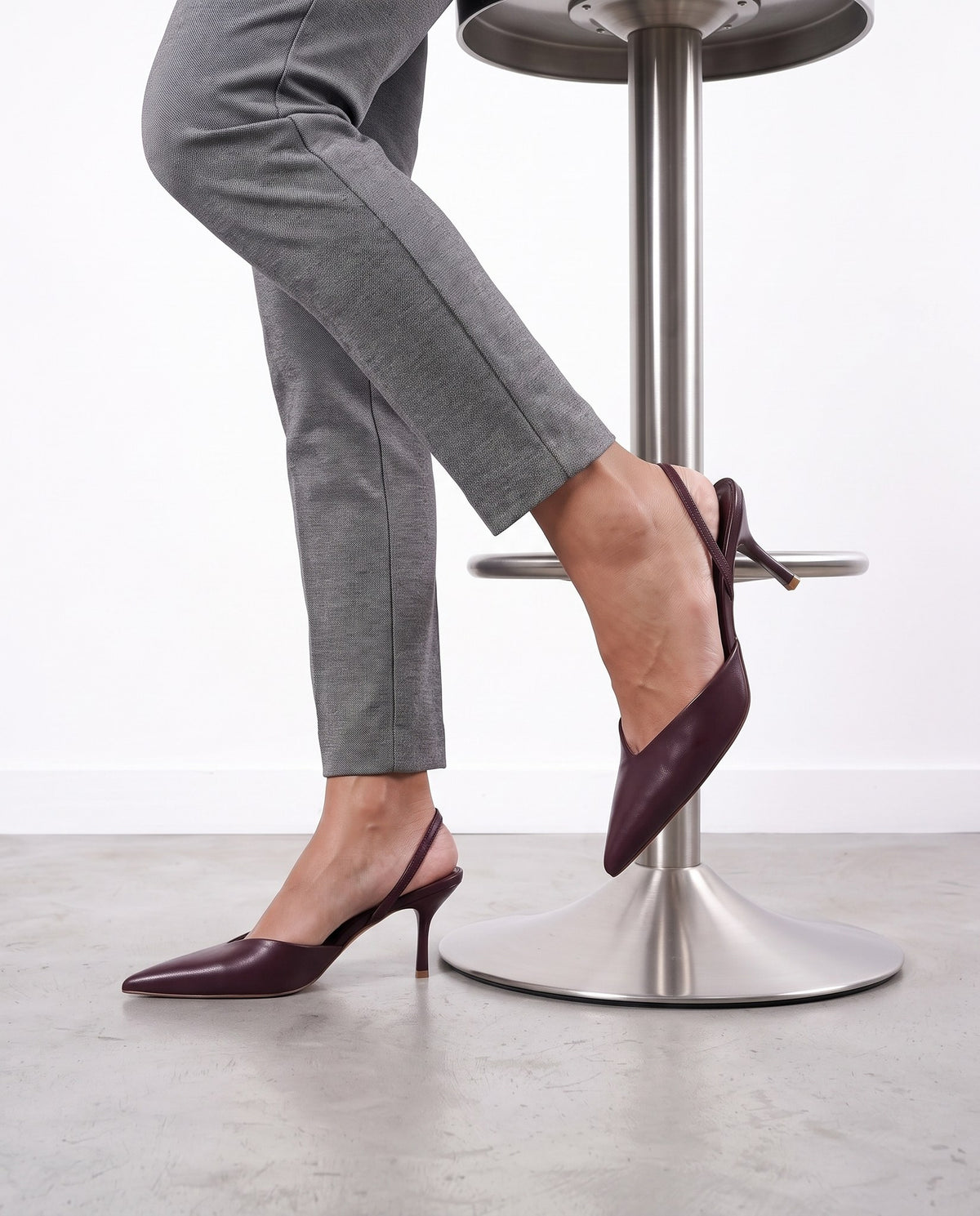 Dettaglio scarpe slingback bordeaux con tacco sottile, una appoggiata a uno sgabello, pavimento in cemento chiaro.
