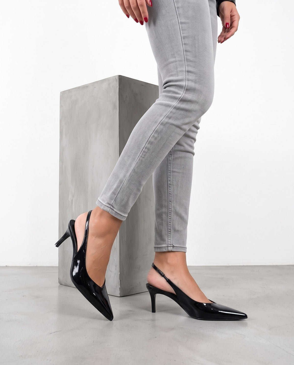 Gambe femminili con décolleté slingback in vernice nera, tacco sottile, accanto a un blocco di cemento