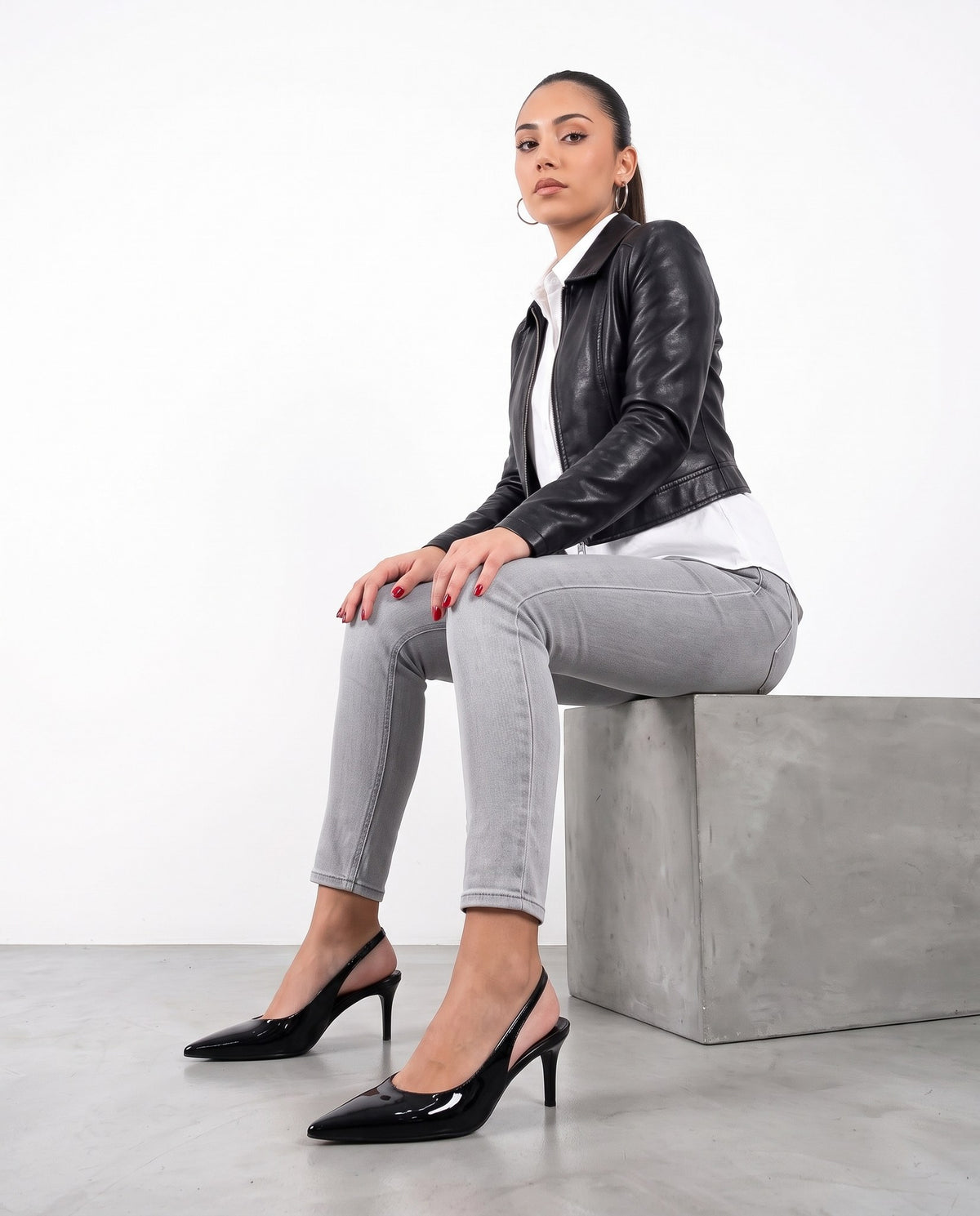 Modella seduta su blocco di cemento, gambe incrociate, décolleté slingback nere in vernice in primo piano
