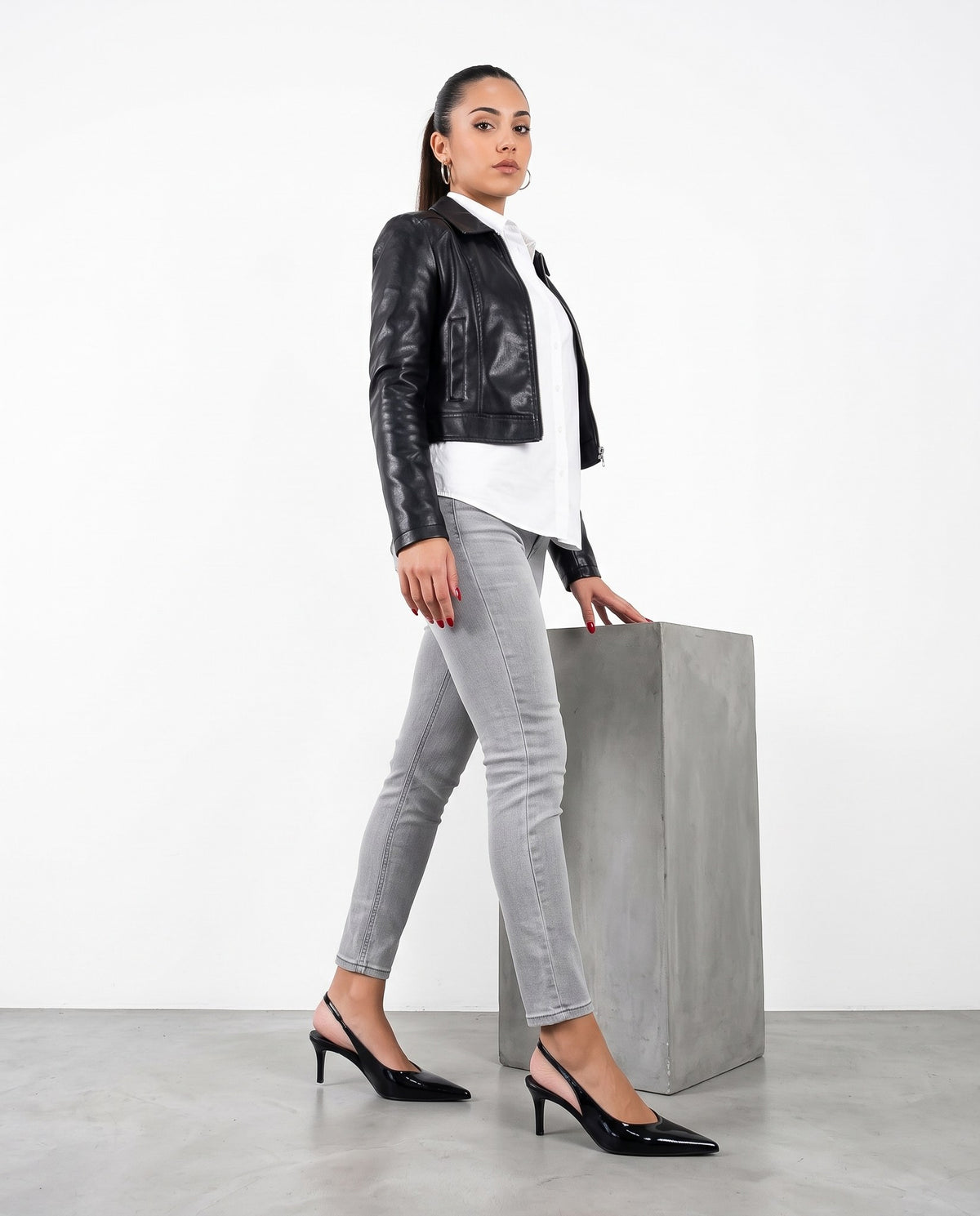 Modella in jeans grigi e giubbino di pelle con décolleté slingback nere lucide, accanto a un blocco in cemento.
