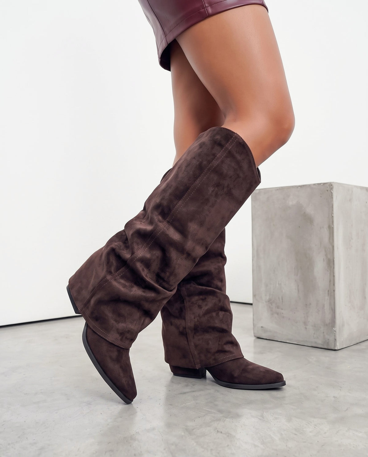 Gambe della modella con stivali scamosciati marroni al ginocchio, accanto a un blocco di cemento grigio