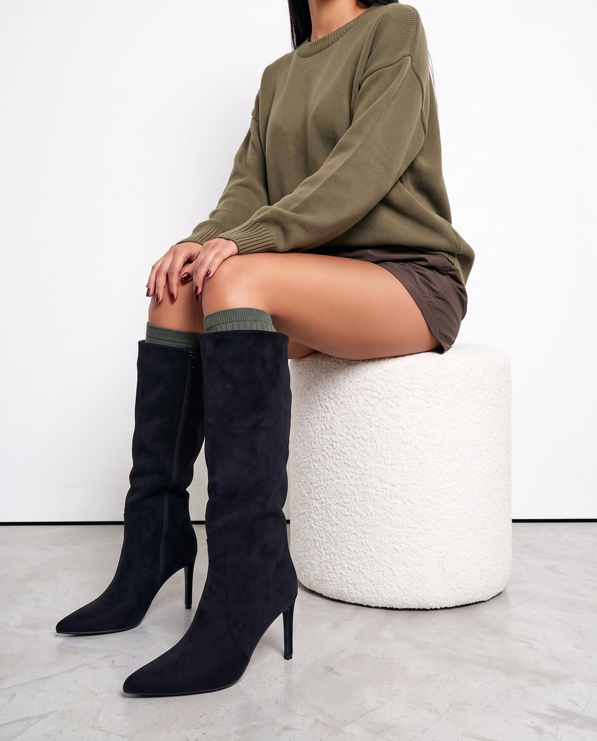 Modella seduta su pouf bianco, stivali neri in camoscio al ginocchio con tacco a stiletto, gambe accavallate.