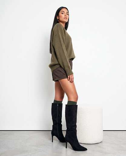 Modella in piedi di profilo con stivali al ginocchio in camoscio nero, maglia oliva e shorts marroni, accanto a pouf bianco