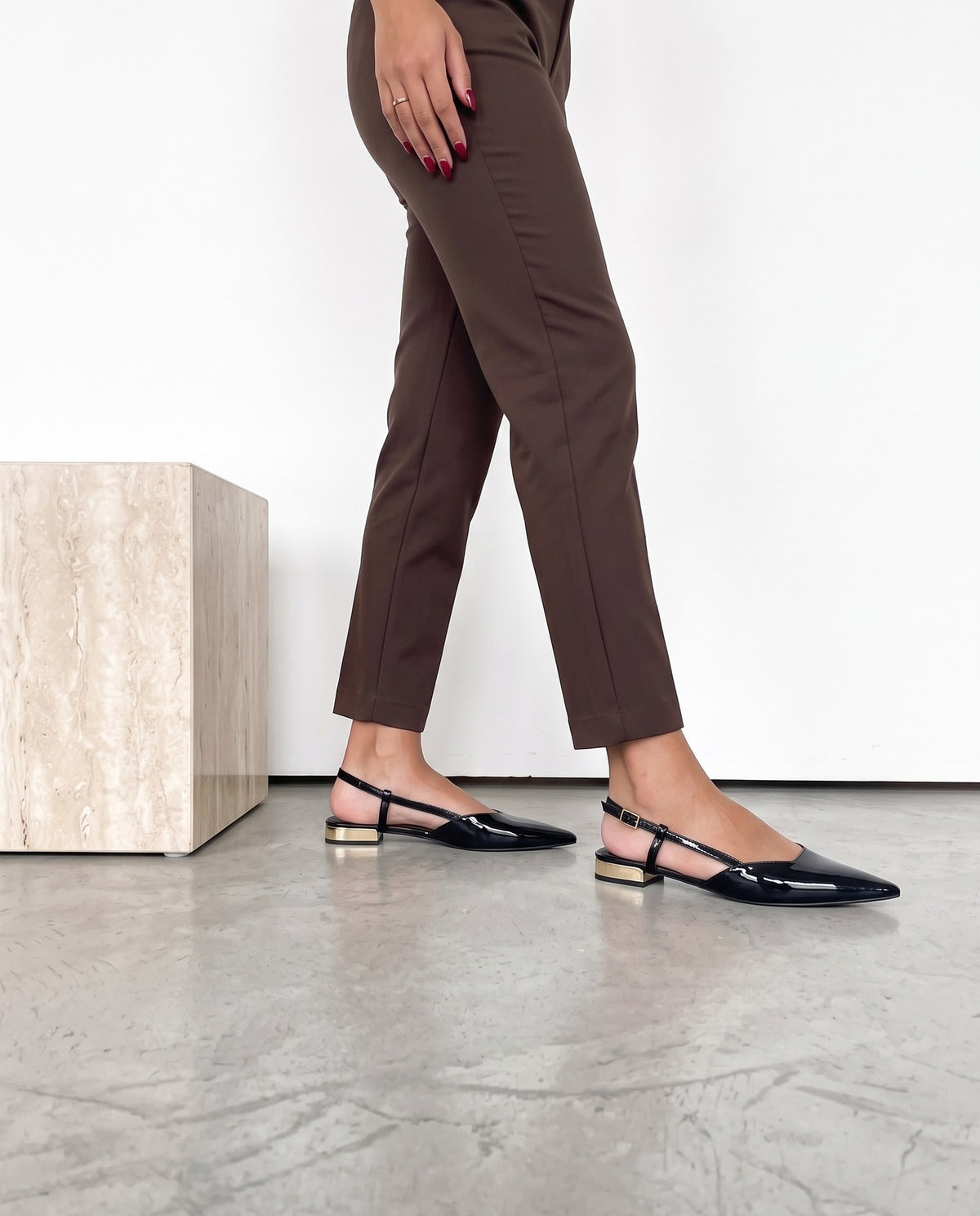 Gambe di modella in piedi con pantaloni cioccolato e slingback nere in vernice con tacco dorato, cubo in travertino accanto.