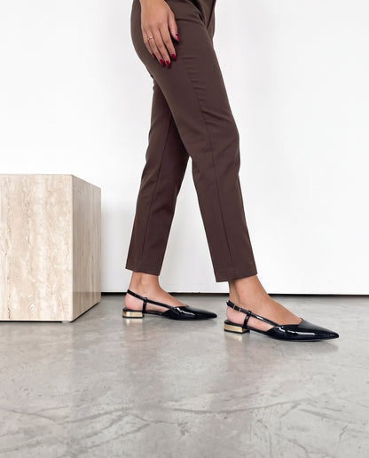 Gambe di modella in piedi con pantaloni cioccolato e slingback nere in vernice con tacco dorato, cubo in travertino accanto.