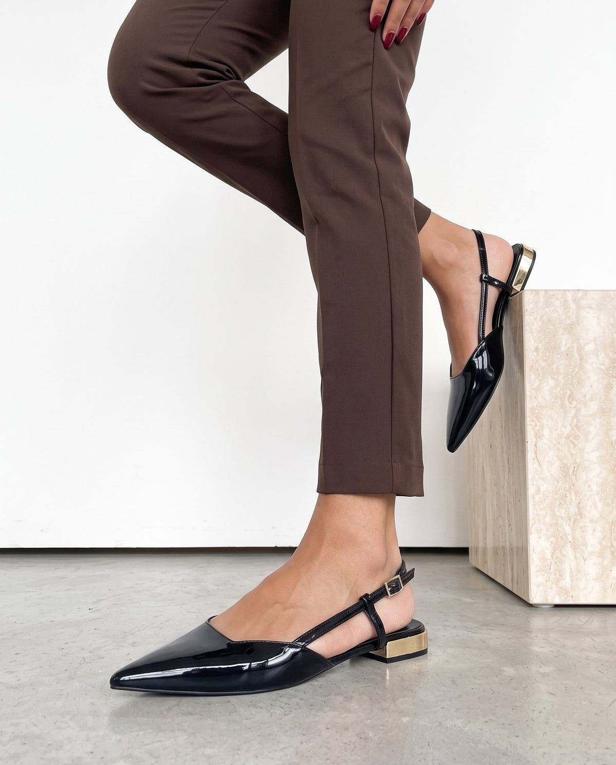 Dettaglio ravvicinato di slingback nere in vernice con tacco dorato, vicino a un cubo in travertino.
