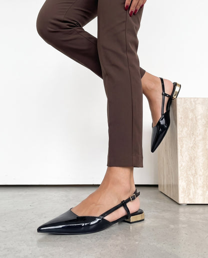 Dettaglio ravvicinato di slingback nere in vernice con tacco dorato, vicino a un cubo in travertino.