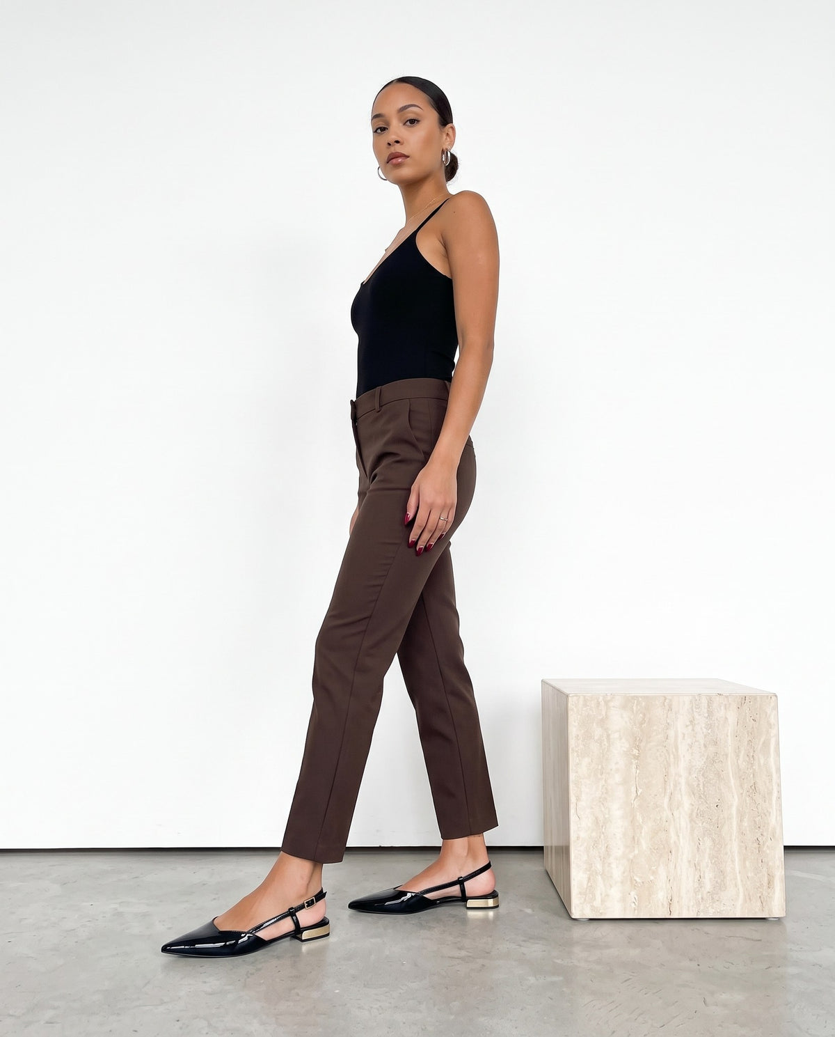 Modella in abito nero e pantaloni cioccolato cammina con slingback nere in vernice, tacco dorato.