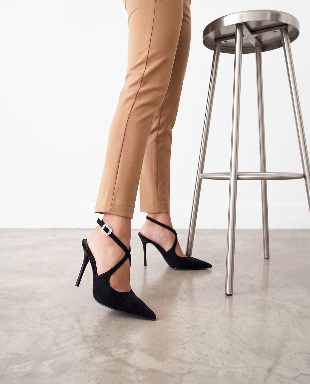 Gambe con pantaloni color cammello e scarpe décolleté nere, accanto a uno sgabello in acciaio.