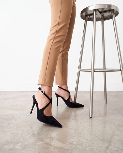 Gambe con pantaloni color cammello e scarpe décolleté nere, accanto a uno sgabello in acciaio.