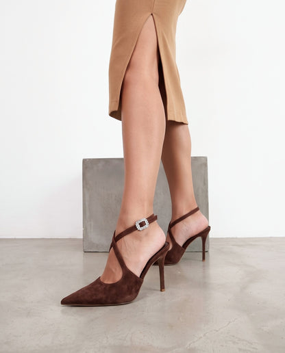 Gambe femminili con scarpe décolleté in camoscio marrone, cinturino incrociato e fibbia gioiello, blocco di cemento sullo sfondo.