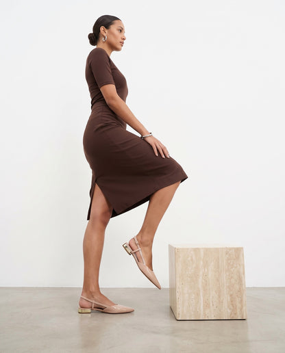 Modella di profilo con abito cioccolato, ballerine slingback rosa nude, cubo in travertino, pavimento in cemento chiaro.