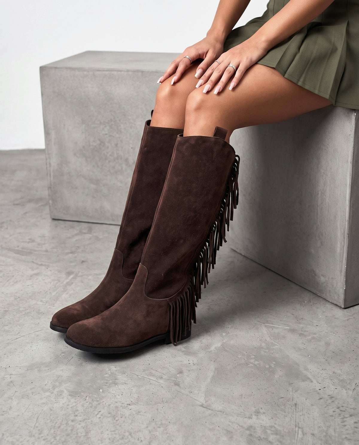 Modella seduta su blocco di cemento, stivali in camoscio marrone testa di moro con frange, gambe accavallate