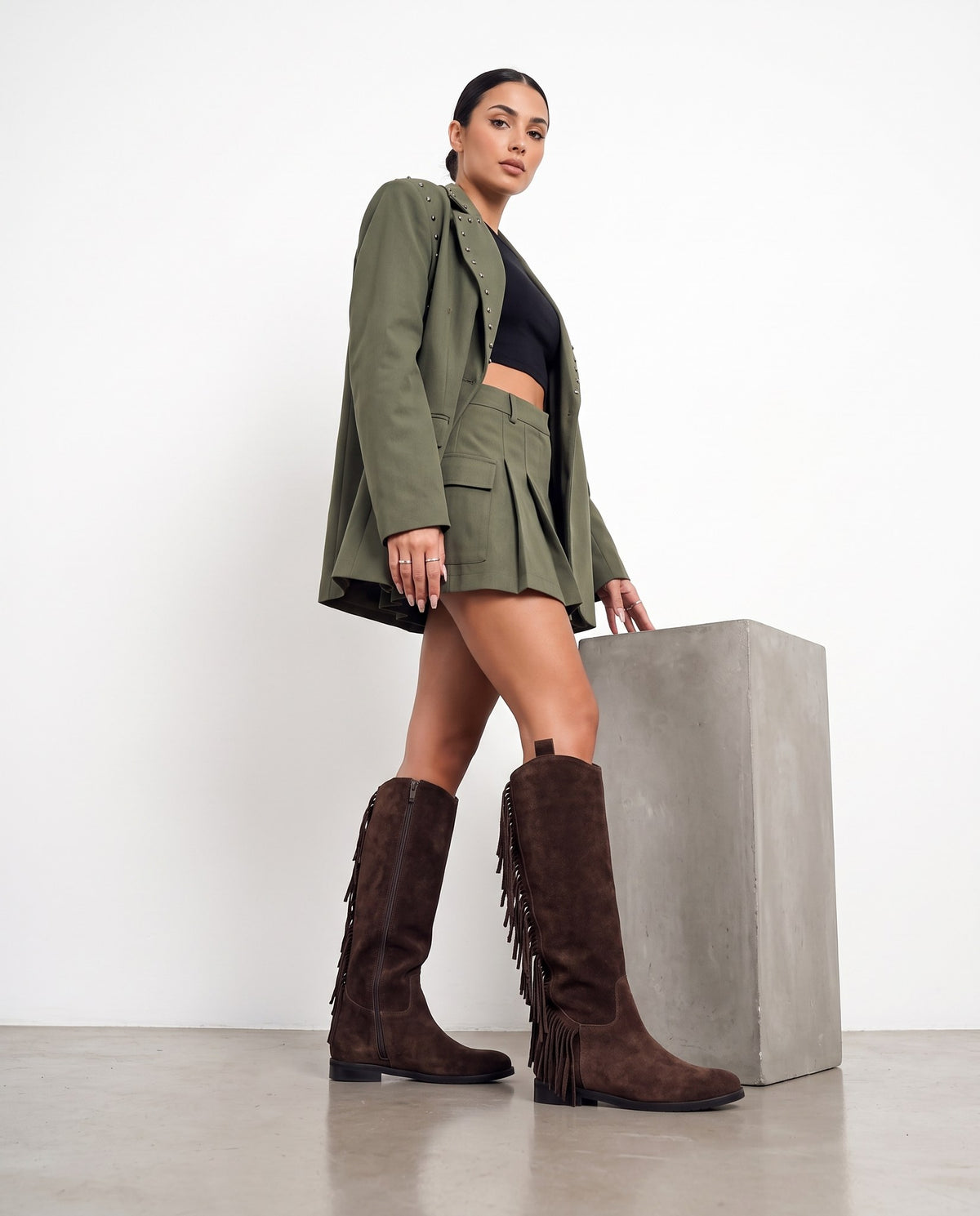 Modella in blazer e minigonna verde militare con stivali camoscio testa di moro con frange, accanto a un blocco di cemento
