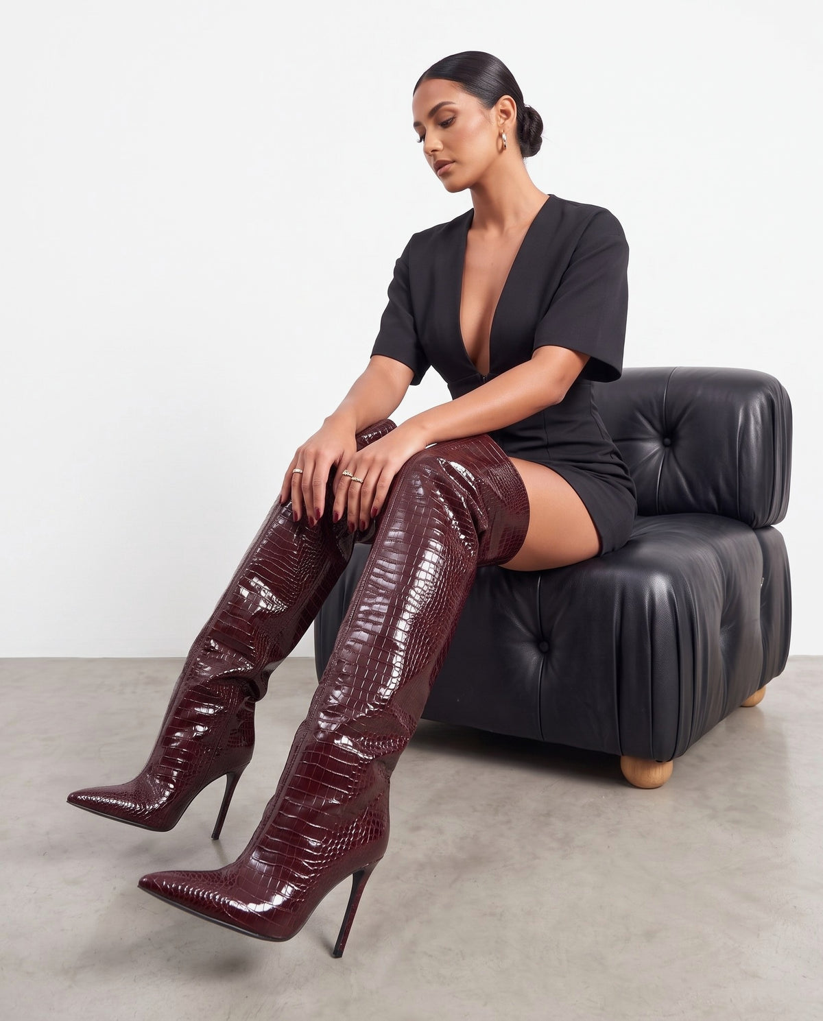 Modella seduta con stivali bordeaux cuissard effetto coccodrillo, abito nero, poltrona in pelle nera