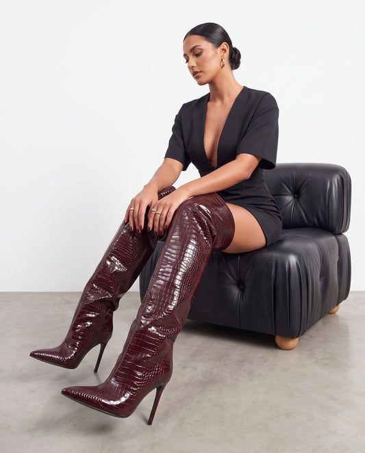 Modella seduta con stivali bordeaux cuissard effetto coccodrillo, abito nero, poltrona in pelle nera