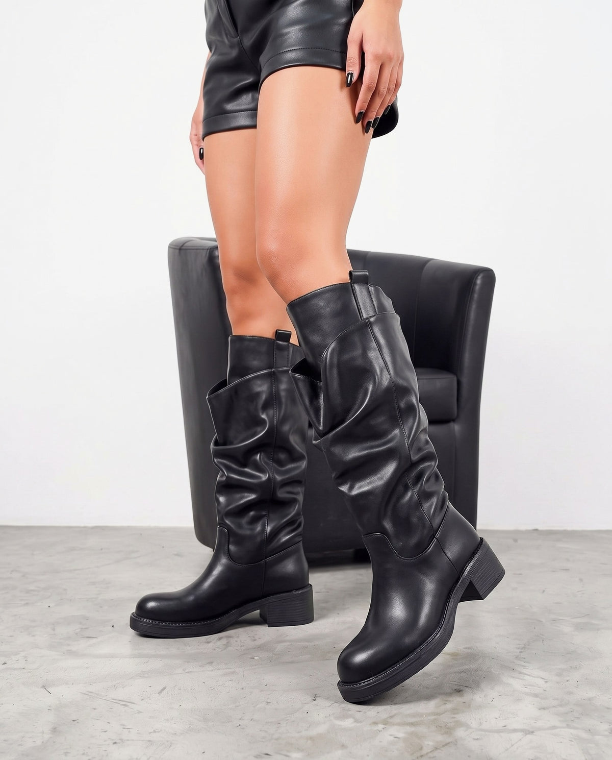 Dettaglio ravvicinato di due stivali neri al ginocchio in ecopelle, con poltroncina nera e pavimento in cemento chiaro.