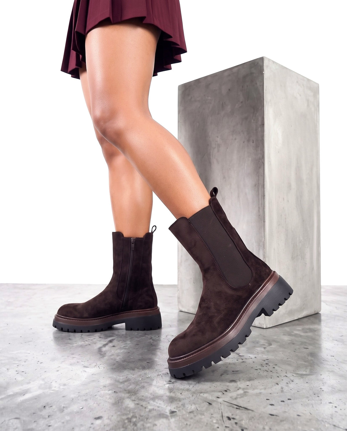 Stivaletti chelsea in camoscio marrone scuro con suola carrarmato, gambe modella in primo piano accanto a blocco in cemento