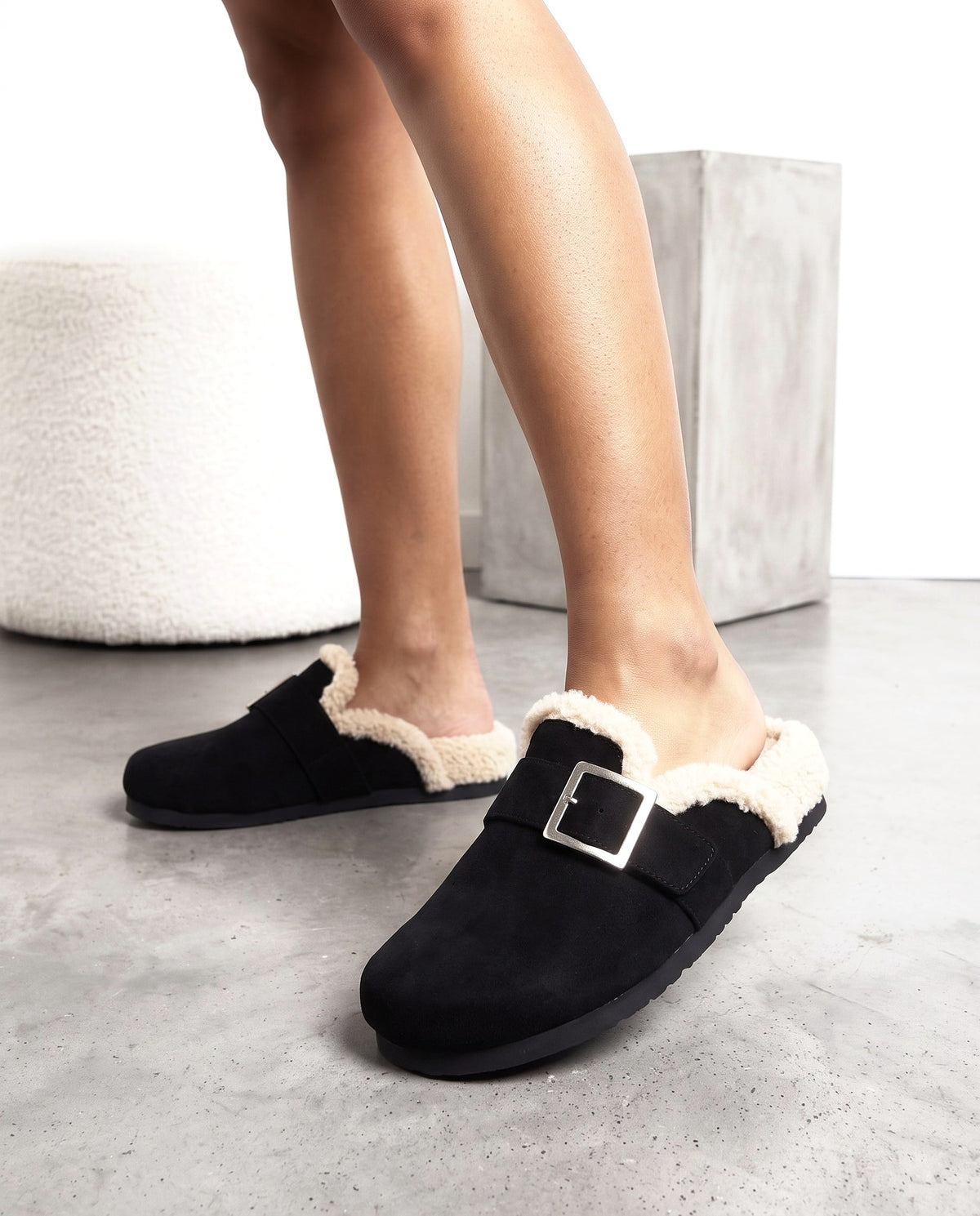 Primo piano delle gambe con ciabatte in camoscio nero con fibbia e bordo in shearling, su pavimento in cemento.