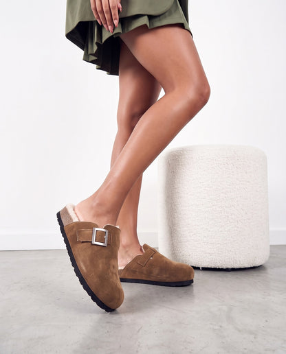 Gambe di modella con pantofole in camoscio marrone e fibbia argento, accanto a pouf bianco bouclé