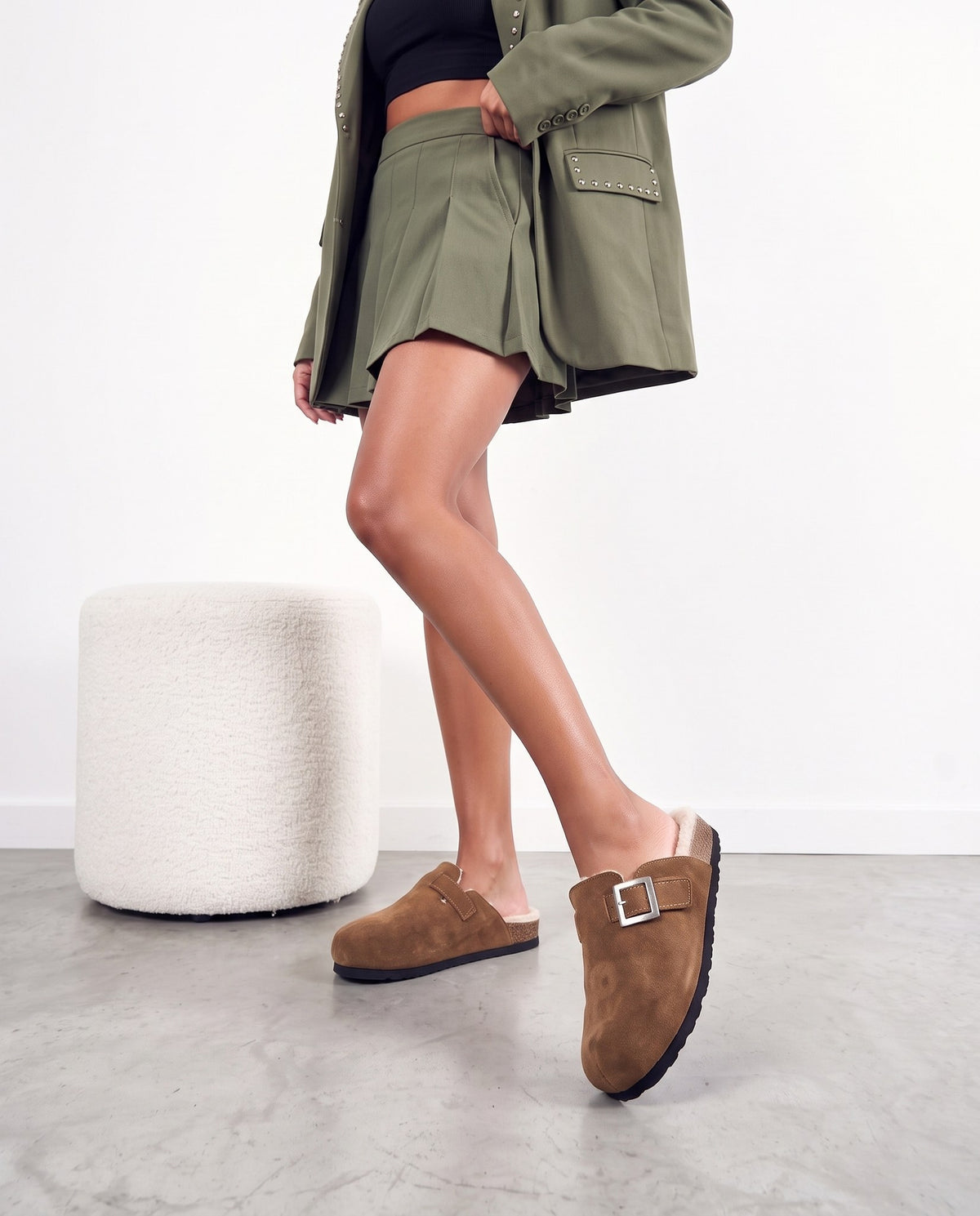 Primo piano di gambe con zoccoli in camoscio marrone, fibbia argento, bordo sherpa panna, su pavimento in cemento chiaro