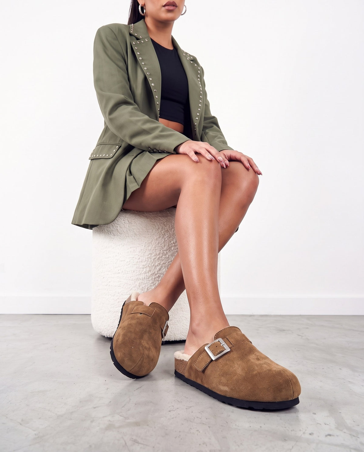 Modella seduta su pouf bianco, gambe accavallate, indossa pantofole mules in camoscio marrone con fibbia argento