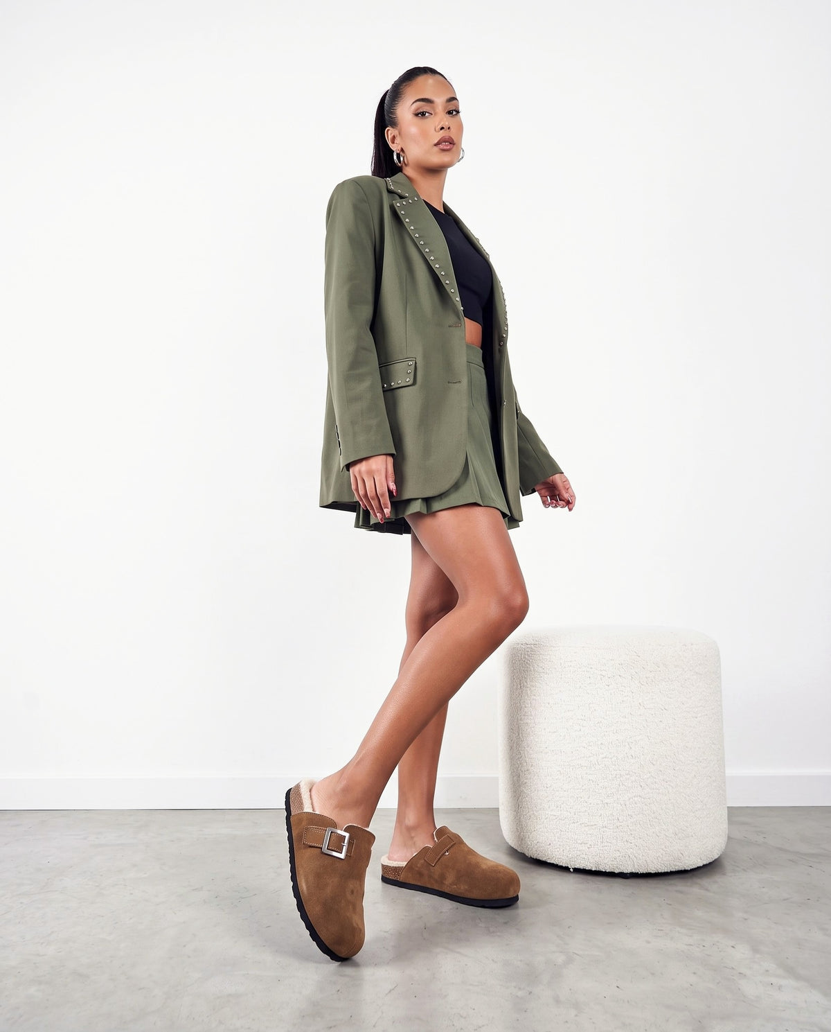 Modella in blazer verde militare e minigonna plissettata indossa pantofole in camoscio marrone con fibbia argento, accanto a un pouf bianco.