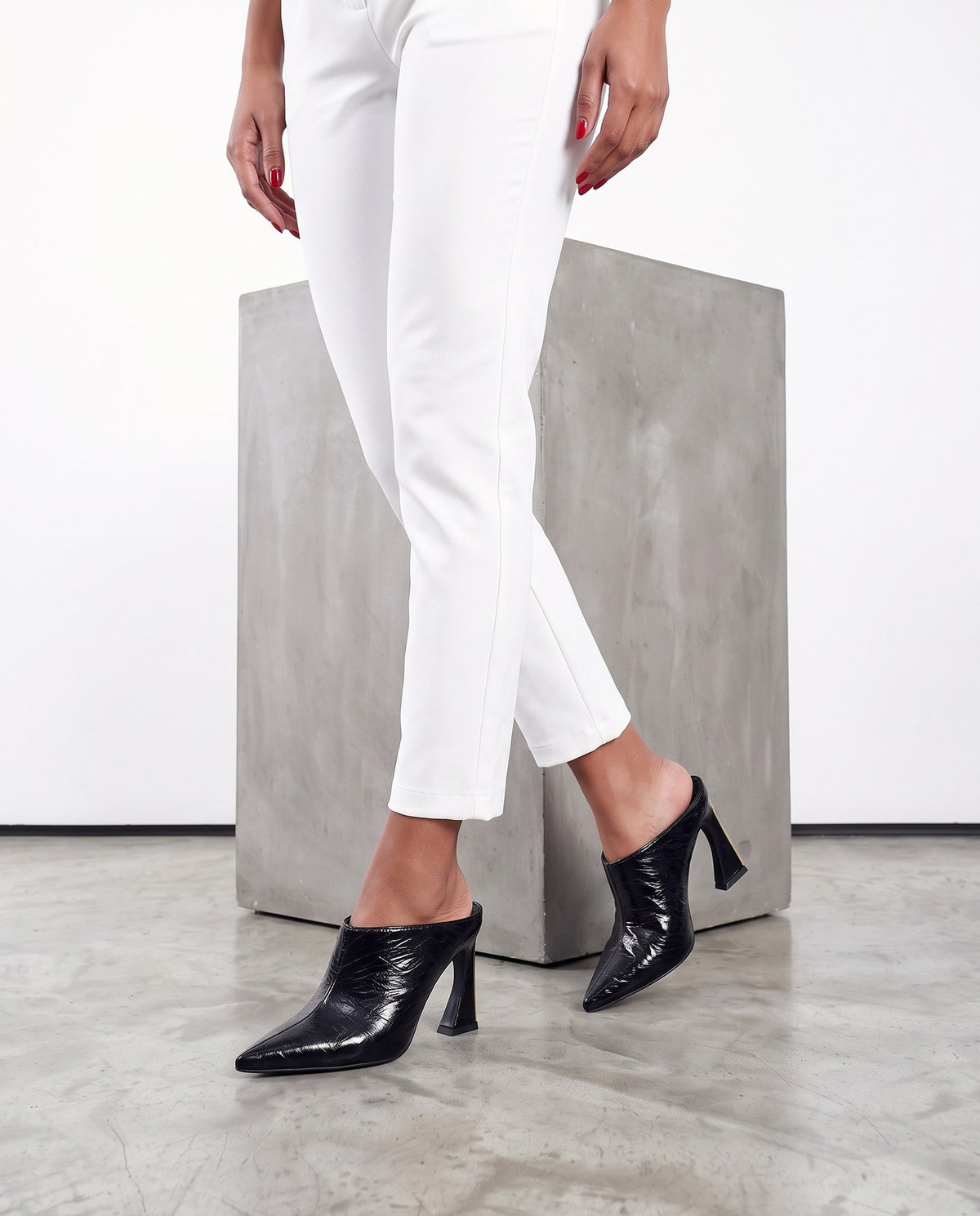 Gambe femminili con mules nere in pelle lucida goffrata, accanto a un blocco di cemento, su pavimento grigio
