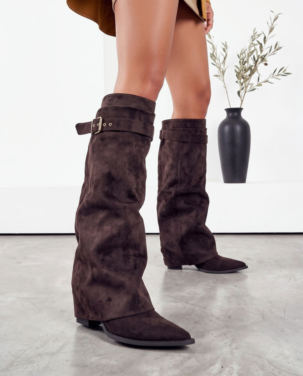 Stivali in camoscio marrone scuro con fibbia, gambe della modella in primo piano, vaso nero con rami sullo sfondo.