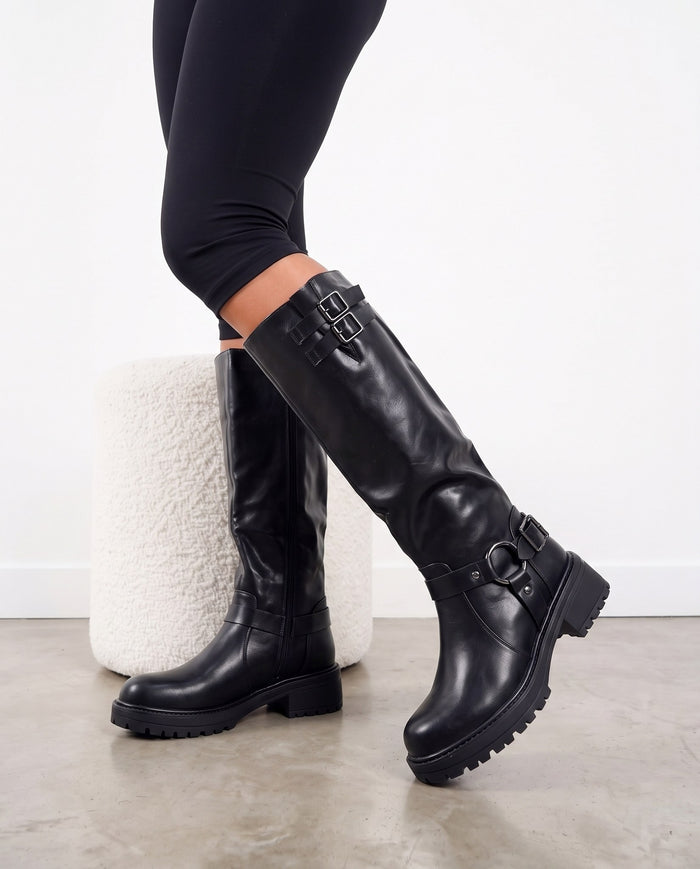 Gambe con stivali alti neri al ginocchio con fibbie, accanto a pouf bianco su pavimento in cemento chiaro