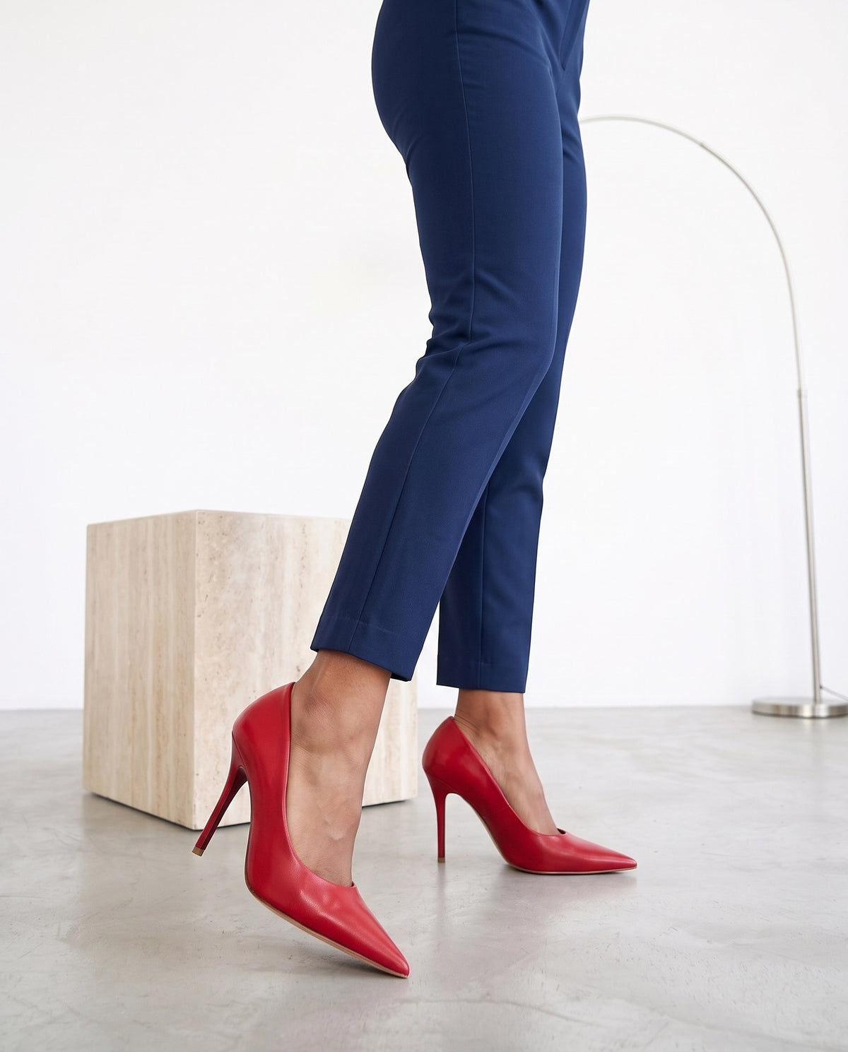 Gambe con pantaloni blu e décolleté rosse, cubo di travertino e lampada ad arco sullo sfondo