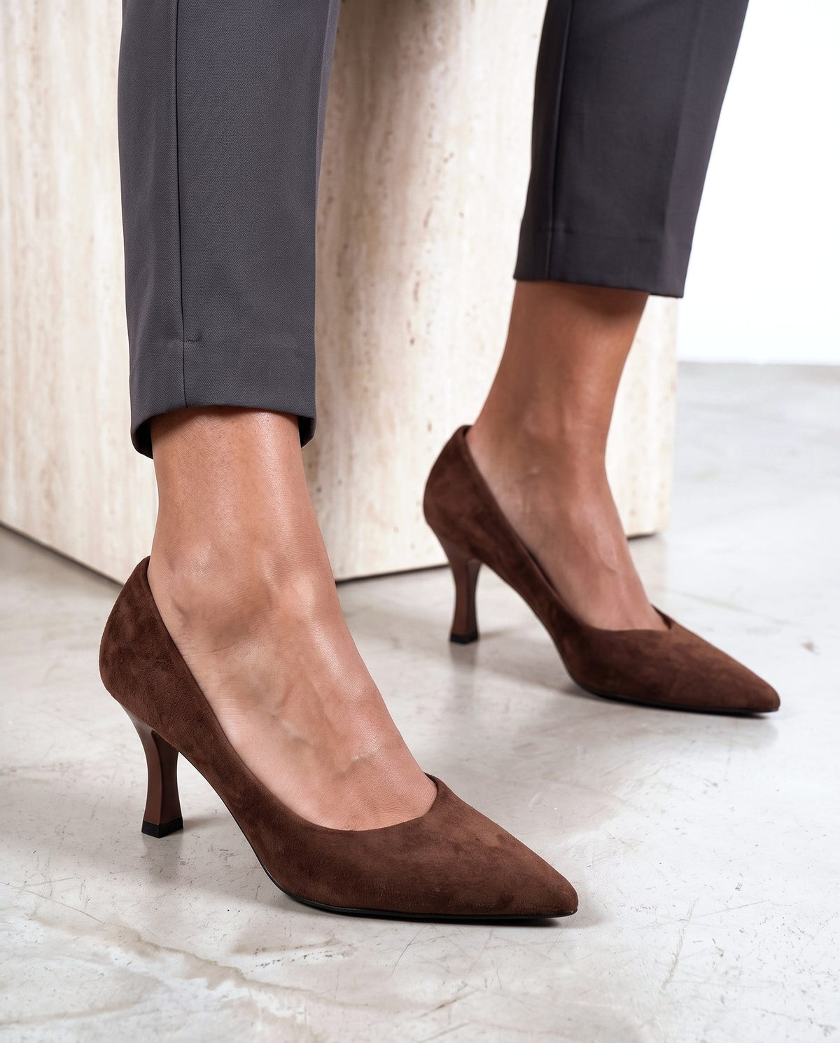 Dettaglio di due scarpe décolleté in camoscio marrone con tacco medio, vicino a cubo in travertino