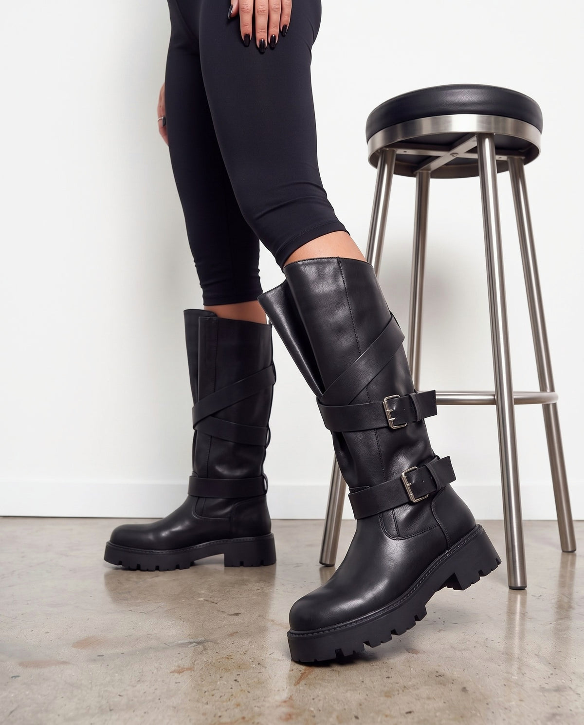 Stivali neri biker con fibbie argento e cinghie incrociate, inquadratura gambe accanto a sgabello